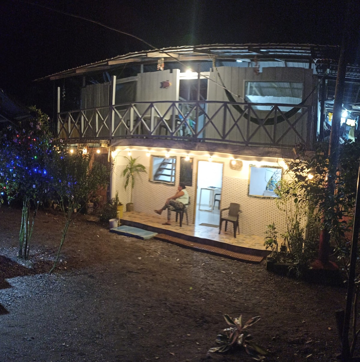 The two-story accommodation is illuminated by warm lights, highlighting a breezy outdoor area with a welcoming porch. A person is seated on the patio, surrounded by lush greenery and decorative plants, creating a relaxed ambiance. The exterior features wooden railings and a tiled entryway.