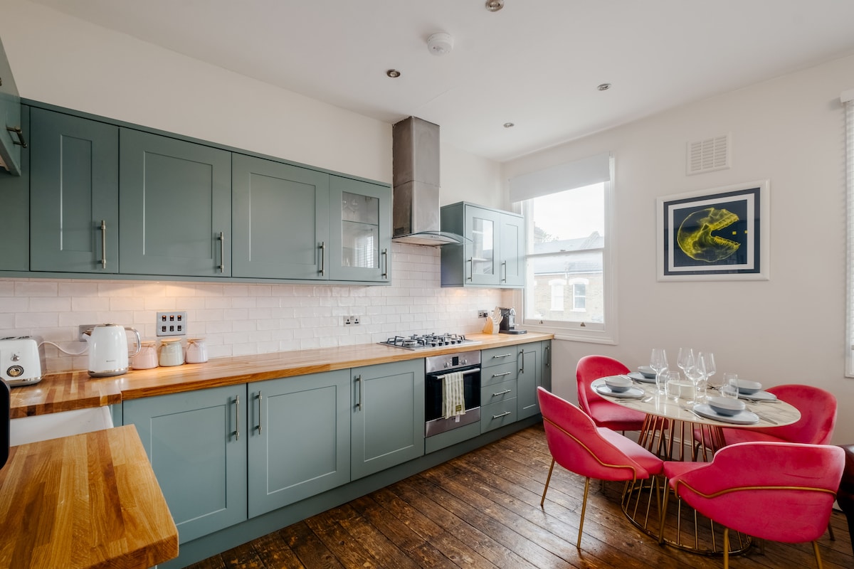 A modern kitchen is highlighted by sleek green cabinetry and a wooden countertop. A round dining table with pink chairs accommodates four, while natural light enters through a large window. Stainless steel appliances and white tile backsplash add a contemporary touch.