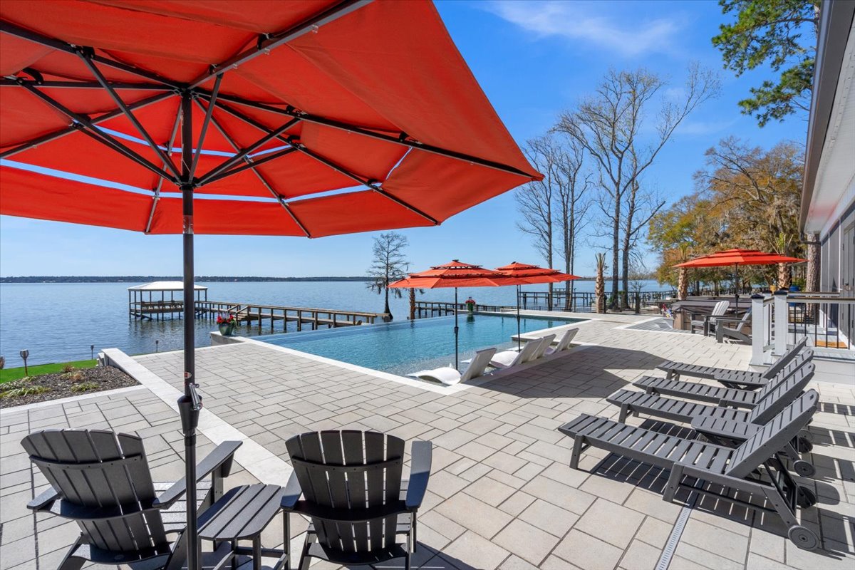 An outdoor patio features lounge chairs and orange umbrellas lining the edge of a stunning infinity pool. The pool's edge meets the serene lake, with a private dock visible in the distance among trees, creating a tranquil retreat for relaxation.
