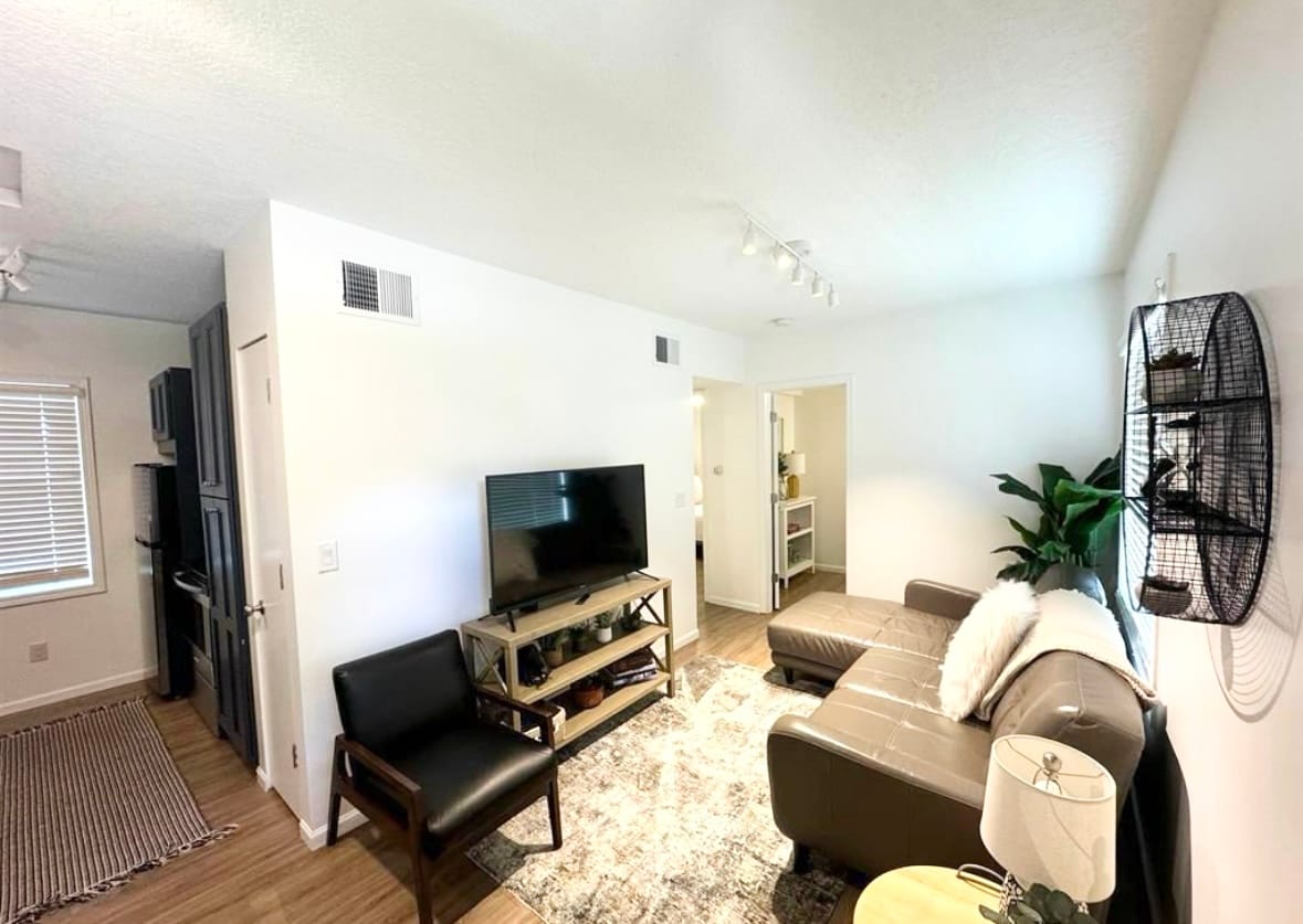 A cozy living area is highlighted by a comfortable sectional sofa and a wooden coffee table. A flat-screen television is mounted on the wall, while a chair adds additional seating. Natural light enters through a nearby window, complementing the minimalist decor.