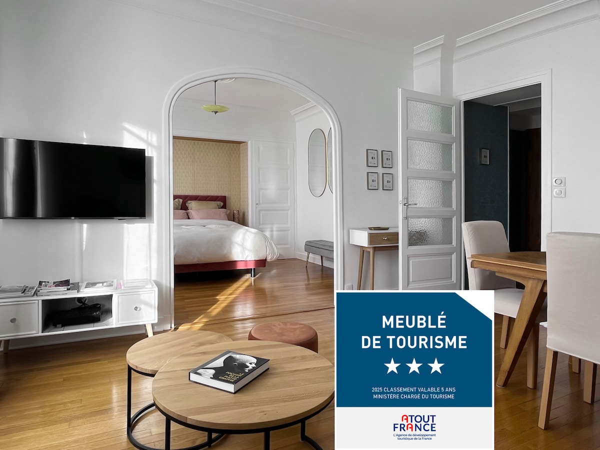 A bright and spacious living area is featured, showcasing a comfortable seating arrangement with a light wooden coffee table. A large TV is mounted on the wall opposite a stylish archway leading to the bedroom, where a spacious bed is discernible in a softly lit environment.