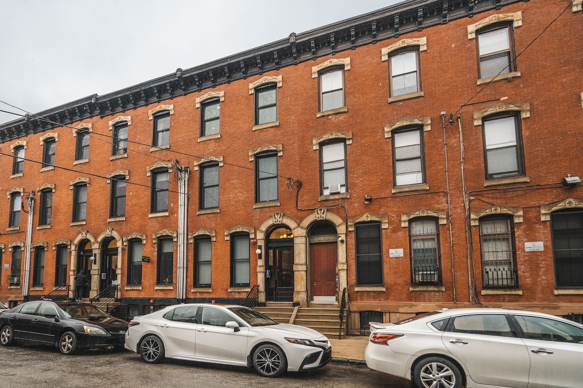 The exterior of a charming brick building is displayed, featuring multiple stories with large windows and decorative elements. The entrance is framed by steps leading to the door. Cars are parked along the street, providing a view of the neighborhood's residential character.