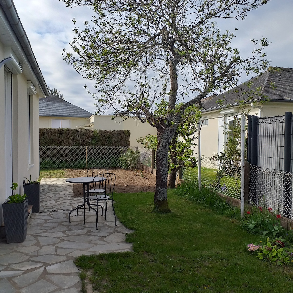 A serene outdoor space features a stone patio with a small round table surrounded by black chairs. A tree provides shade, complemented by greenery and blooming flowers along the edge. A fenced area and a garden can be seen in the background, adding to the tranquil atmosphere.