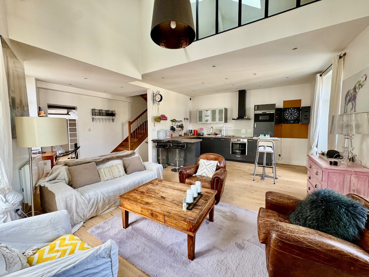 The open-plan living area features high ceilings and abundant natural light. A wooden coffee table is centered on a soft area rug, surrounded by a mix of comfortable seating. The kitchen is equipped with modern appliances, and a staircase leads to the upper level.