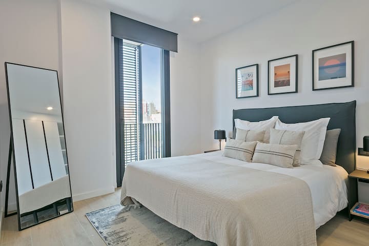 Светла и спокойна спалня с удобно двойно легло, чисти бели чаршафи и декоративни възглавници. Голям прозорец с щори внася естествена светлина. Минималистичният декор включва рамкирани произведения на изкуството, нощни шкафчета с лампи и огледало в цял ръст.