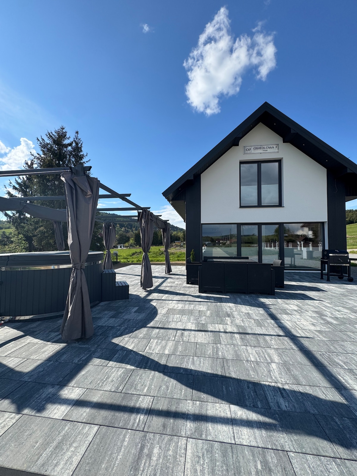 An outdoor patio area is presented with a hot tub and spacious seating arrangements. Shade is provided by draped curtains, and large windows in the house allow for ample natural light. Lush greenery can be seen in the background, complementing the clear blue sky.
