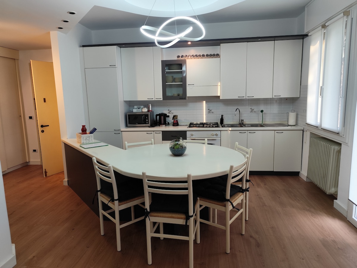 A modern kitchen is equipped with white cabinetry and stainless steel appliances. A large circular dining table is surrounded by light wood chairs. Natural light floods the space through large windows, highlighting the contemporary light fixture overhead.