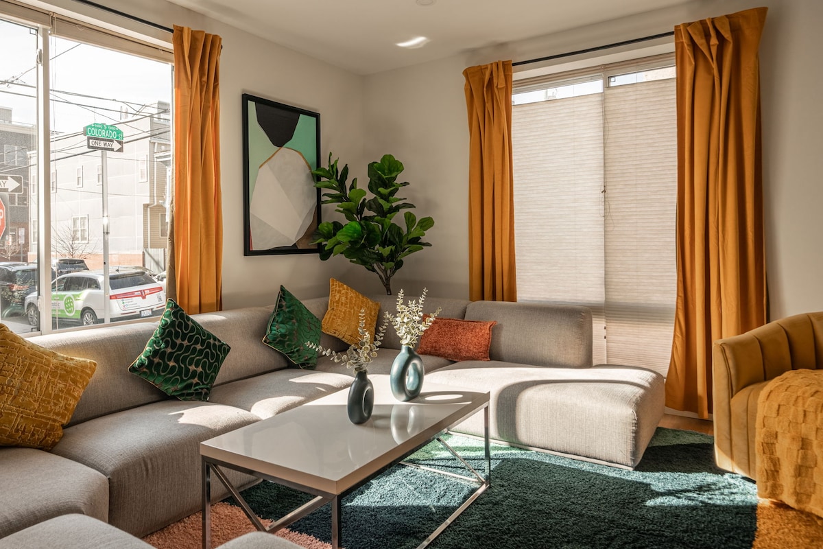 A modern living room features a sectional sofa adorned with colorful cushions, complemented by a coffee table with decorative vases. Large windows allow natural light to brighten the space, while warm orange curtains frame the view. A potted plant adds a touch of greenery.
