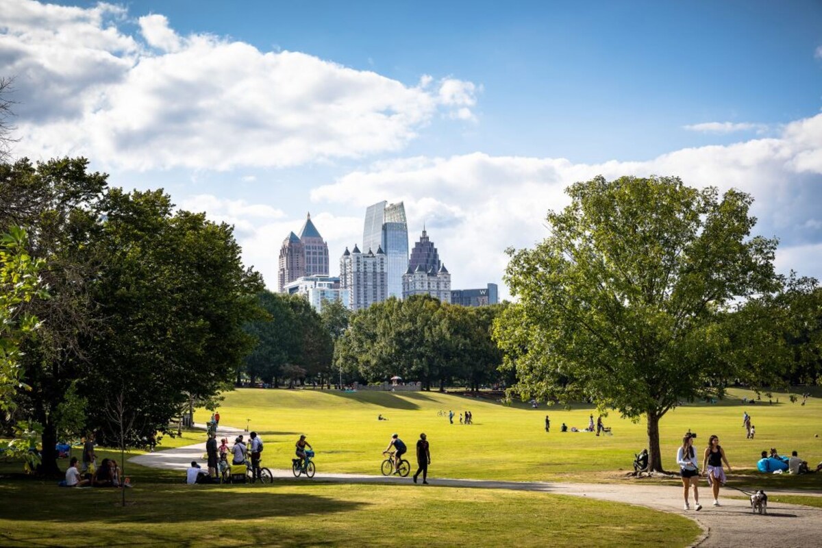 A sprawling green park displays a vibrant landscape filled with trees and shrubs. People can be seen walking and biking along paved paths. The skyline of modern buildings rises in the background under a partly cloudy blue sky.