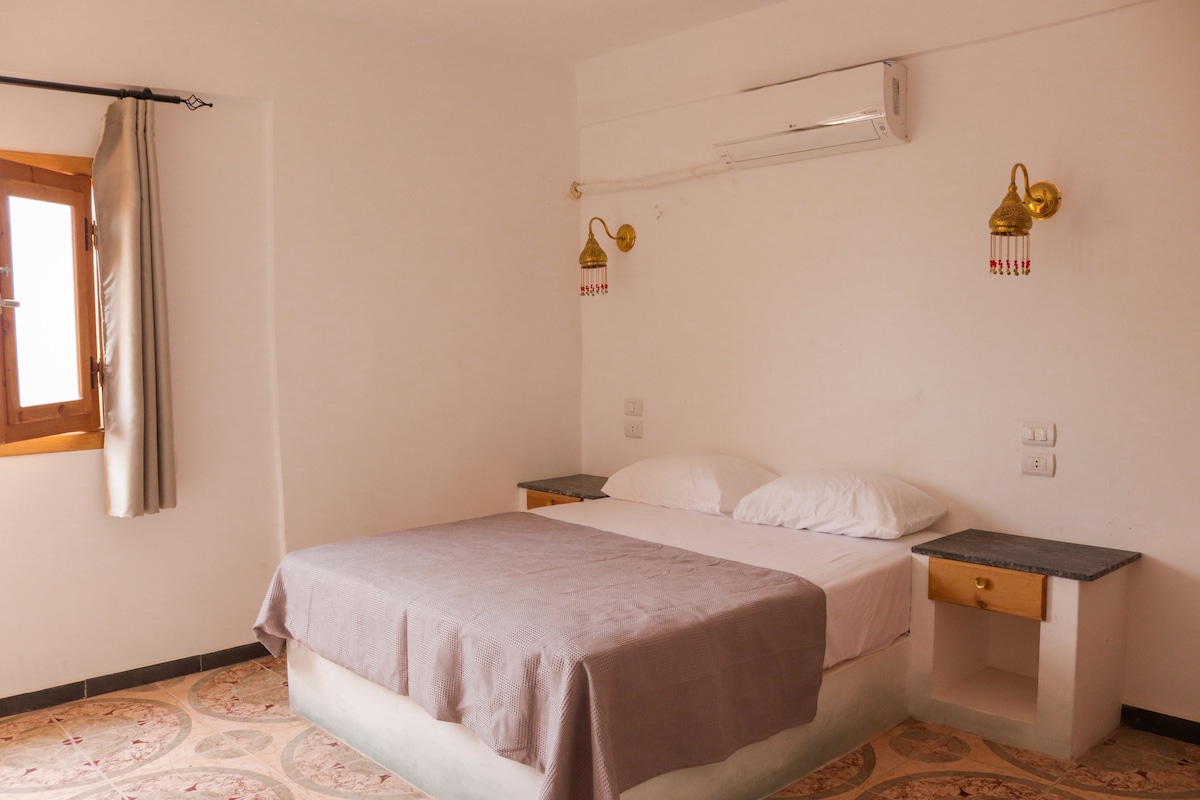 A simple and clean bedroom features a spacious double bed dressed in a light-colored blanket. Two wall-mounted lamps illuminate the room, while a window with curtains allows natural light to enter. A pair of nightstands are positioned on either side of the bed.