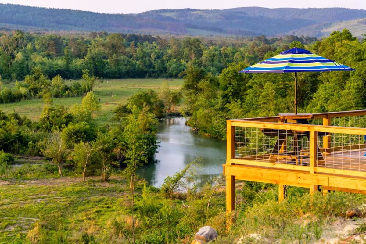 A wooden deck overlooks a tranquil river, surrounded by lush greenery and rolling hills. A striped umbrella provides shade over a small table, creating an inviting outdoor space. The scene is accented by the natural beauty of the mountains in the background.