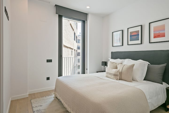 Chumba cha kulala chenye mwanga na kitanda cha watu wawili, matandiko ya kawaida na mchoro ulio juu ya ubao wa kichwa. Dirisha kubwa huleta mwanga wa asili, likionyesha muundo safi, sakafu nyepesi ya mbao na mazingira tulivu, ya kisasa.