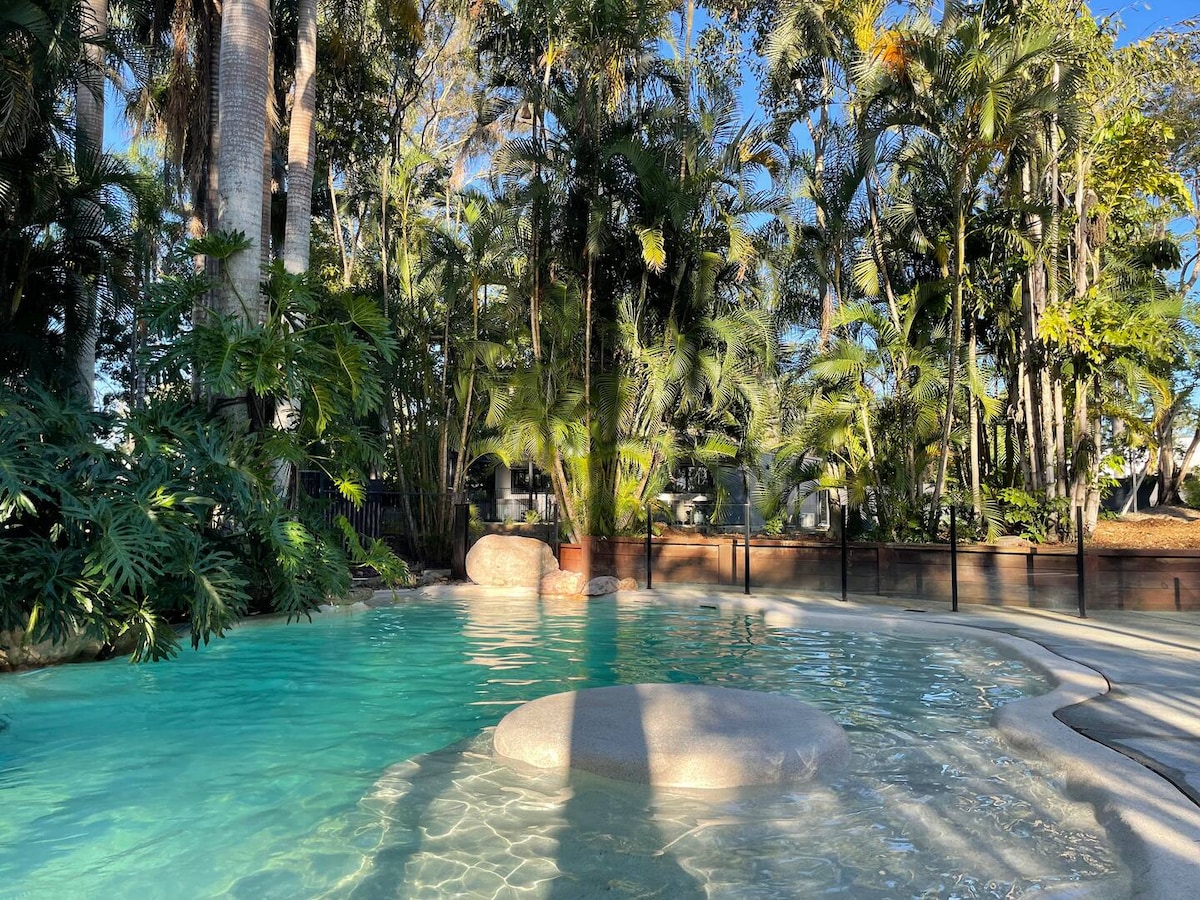 A serene swimming pool is surrounded by lush tropical greenery, featuring smooth stones partially submerged in the water. Sunlight reflects off the shimmering surface, creating a calming atmosphere. The area is designed for relaxation, with a natural feel enhanced by the foliage and light.
