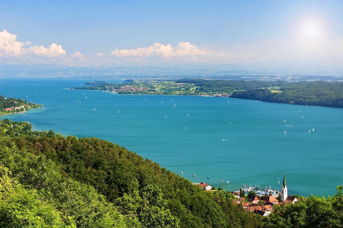 A panoramic view of Lake Constance is displayed, showing a vast expanse of blue water dotted with sailboats. The shoreline features greenery and a small village with a church steeple, framed by mountains in the distance under a clear sky.