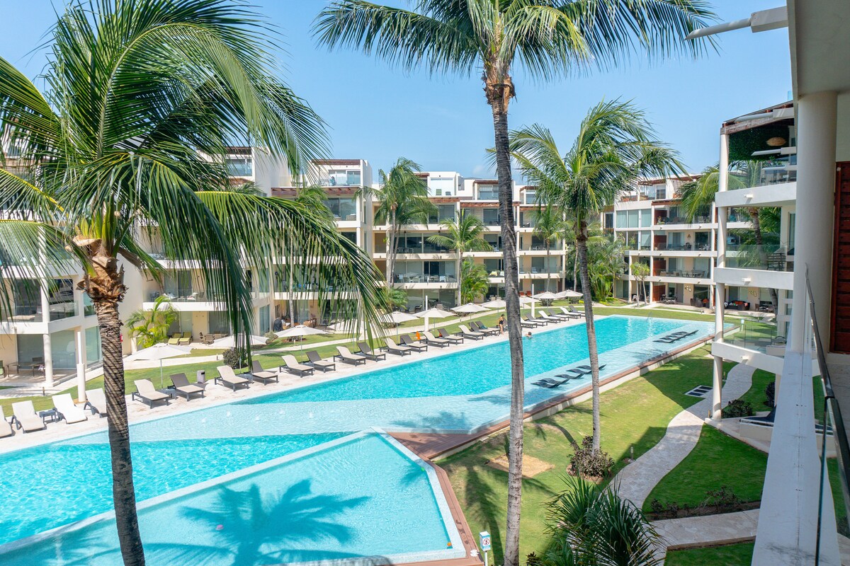 A clear view of a rectangular swimming pool is framed by lush palm trees. The pool is surrounded by lounge chairs and leads to landscaped gardens. Multiple levels of modern accommodations are visible, featuring glass balconies that overlook the pool area.