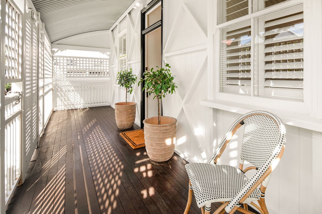 An inviting front verandah features polished wooden flooring and white-paneled walls. Two potted plants add greenery, while natural light creates playful shadows. A single chair with a patterned design sits beside the entrance, offering a comfortable space for relaxation.