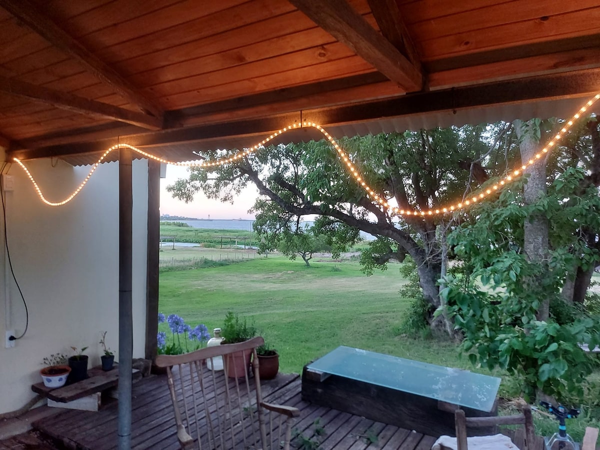 A cozy seating area is seen under a wooden roof, with soft lighting strung above. A wooden bench and a table are positioned on the deck, surrounded by lush greenery and a view of the expansive landscape beyond.