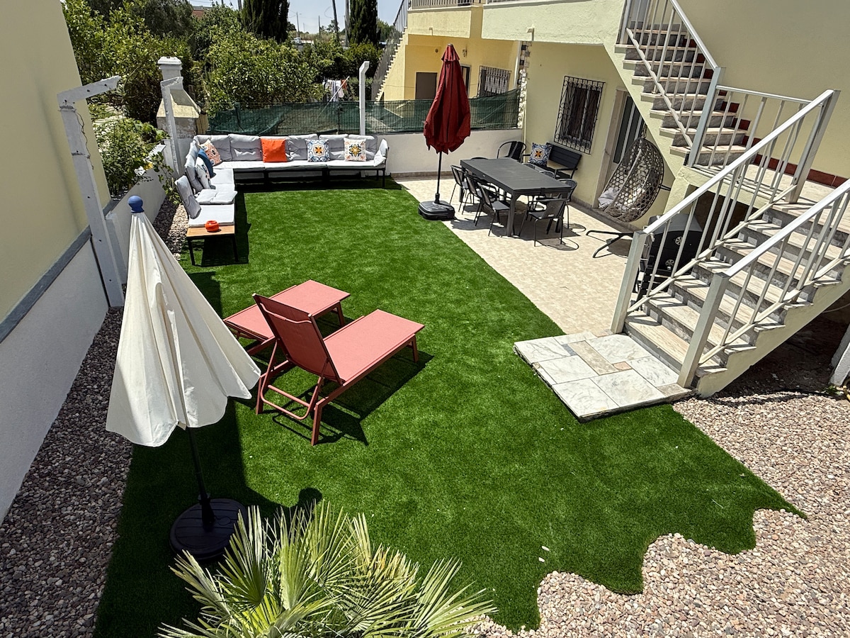 The outdoor space features a well-kept artificial grass area with elegant lounge chairs and a dining table under a shaded umbrella. A comfortable seating arrangement can be seen against the backdrop of a vibrant garden, with stairs leading to the upper level of the house.