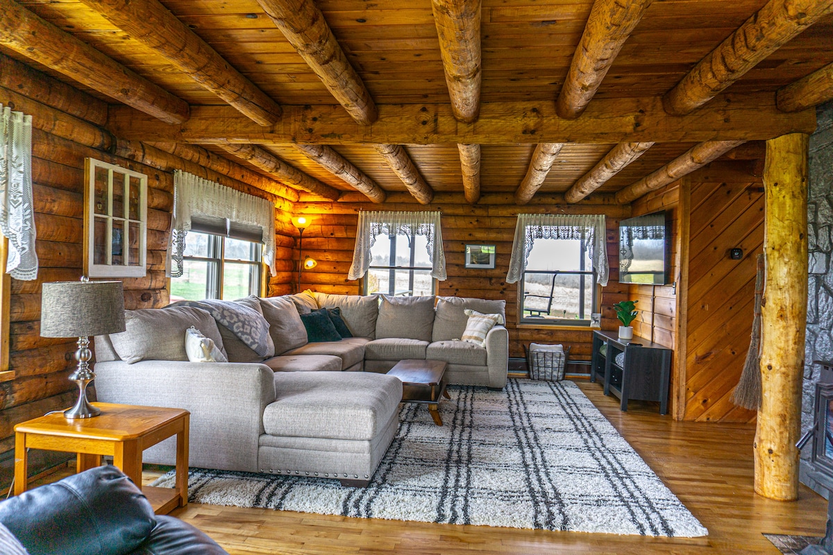 A cozy living room features a large sectional couch arranged around a patterned rug on hardwood flooring. Wooden beams are visible on the ceiling, and windows offer views of the surrounding landscape. A lamp and side table add to the comfortable atmosphere.