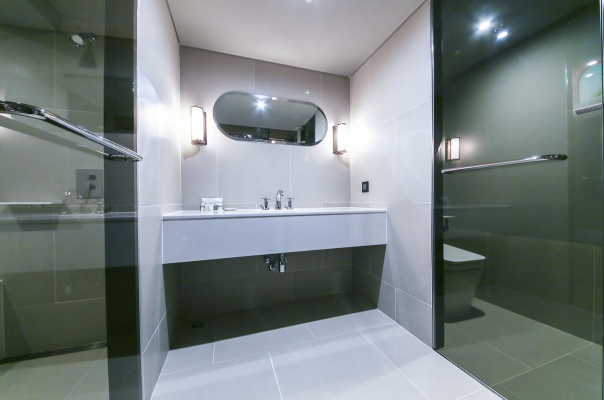 A modern bathroom features sleek gray tiles and a spacious vanity with dual sinks. Ambient lighting is provided by wall sconces beside a large mirror. A minimalist shower area and a discreet toilet area are also included, providing a clean and functional space.
