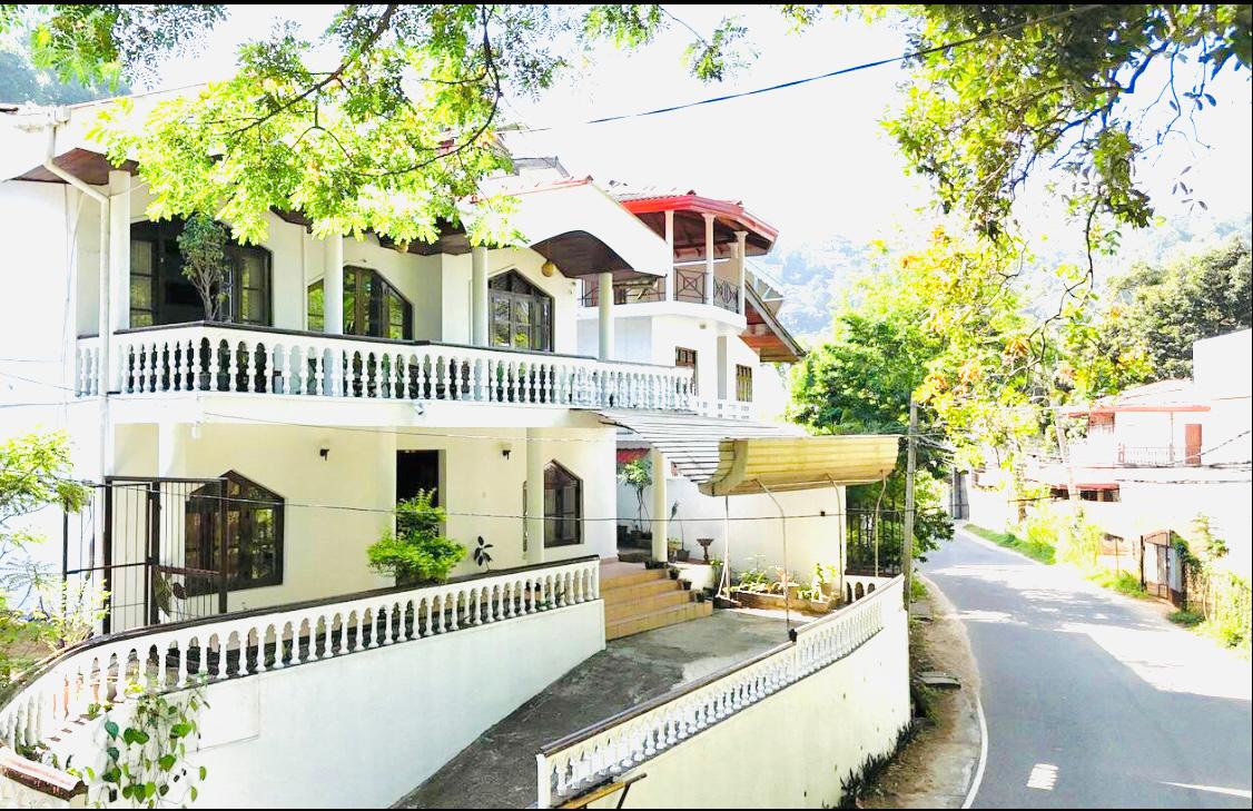 A multi-story bungalow is showcased amidst lush greenery and a clear sky. The exterior is characterized by white walls and decorative balcony railings. A curved driveway leads up to the entrance, while a nearby road is visible, framed by trees and surrounding structures.