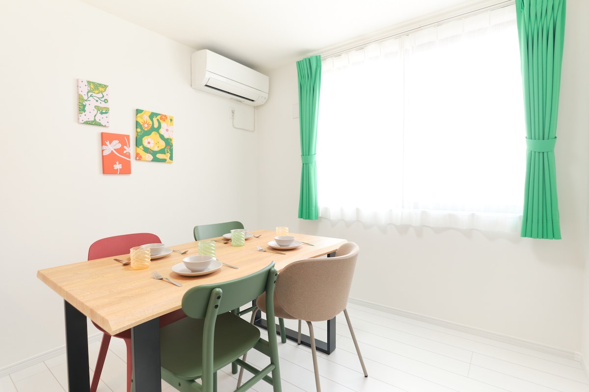 A dining area featuring a wooden table surrounded by four chairs in various colors. Light-colored walls and a window with sheer white curtains allow natural sunlight to illuminate the space. Colorful wall art adds a playful touch to the room.