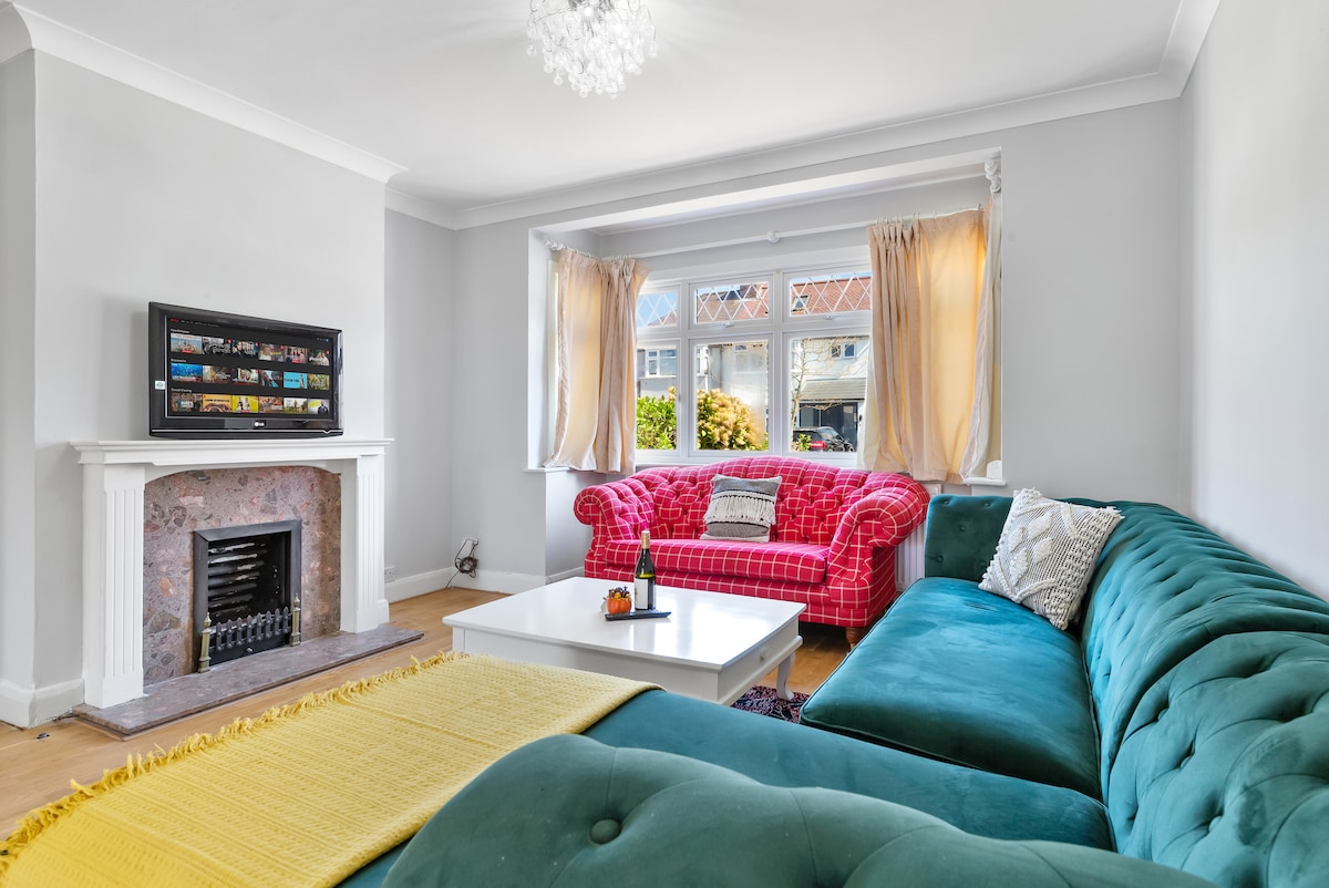 A spacious living area features a large green sectional sofa and a traditional red checkered couch, inviting relaxation. A white coffee table sits at the center, and natural light streams through large windows, highlighting the pale gray walls and cozy fireplace.