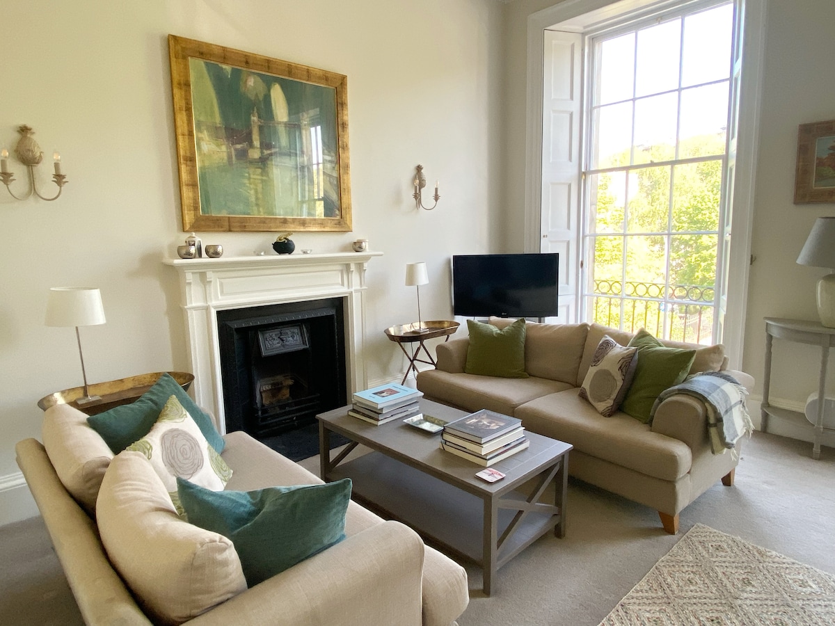 A well-furnished sitting room features two comfortable sofas adorned with decorative pillows. A coffee table sits in front, stacked with books. A marble fireplace and a wall art piece are seen, with large windows allowing natural light and a view of greenery.