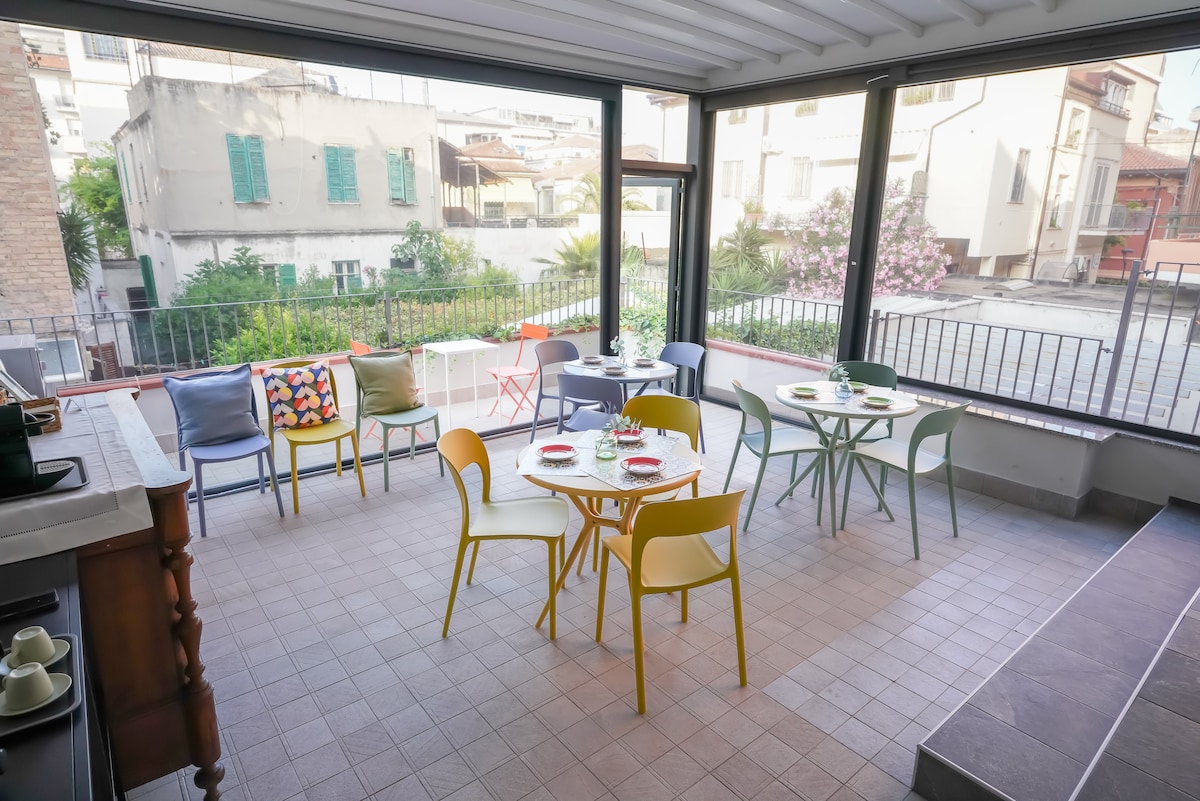 An inviting covered terrace features a mix of dining tables and colorful chairs. Large windows allow natural light to fill the space while showcasing views of the surrounding area. A relaxed atmosphere is created with decorative cushions and potted plants strategically placed throughout.