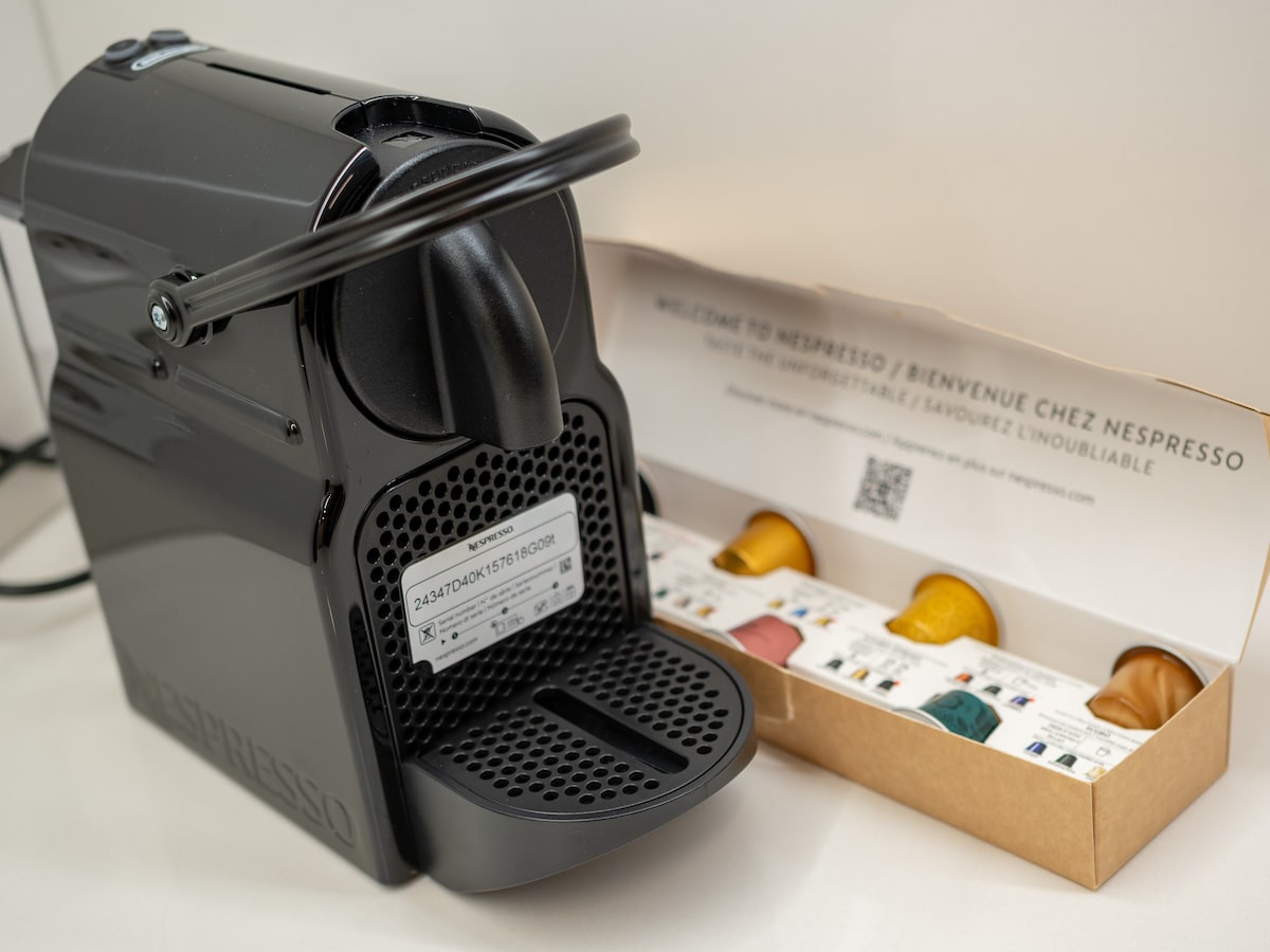 A black Nespresso coffee machine is positioned on a countertop. Next to it, a box containing an assortment of coffee capsules in various colors is displayed, with a clear layout showing the capsule selections.