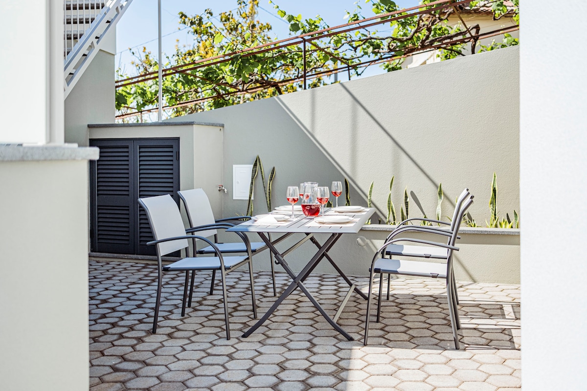 A private outdoor patio features a dining table set for four, with glasses of red wine arranged on white plates. Four modern chairs are positioned around the table, while greenery is visible in the background, providing a relaxed atmosphere for outdoor meals.
