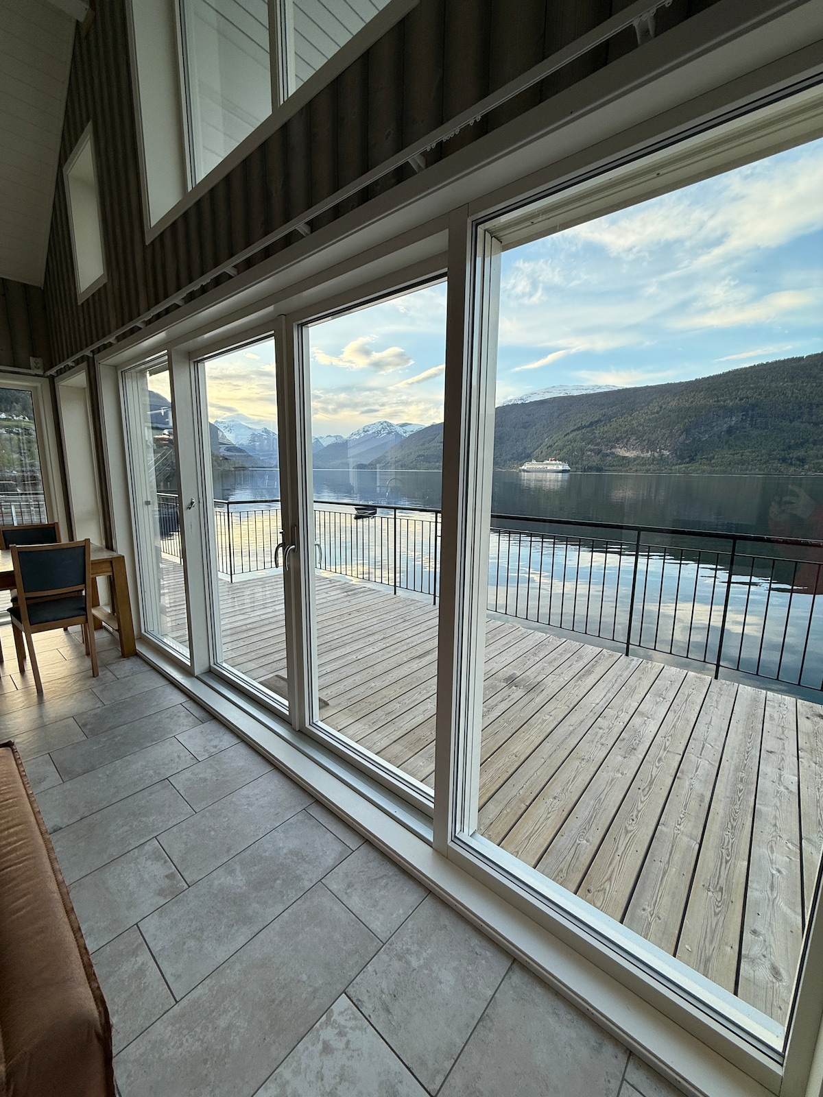 Large glass doors provide a view of the expansive wooden terrace and the serene waters beyond. The surrounding landscape features mountains and a tranquil fjord, with sunlight reflecting off the water, creating a peaceful atmosphere. A seating area is visible inside the space.