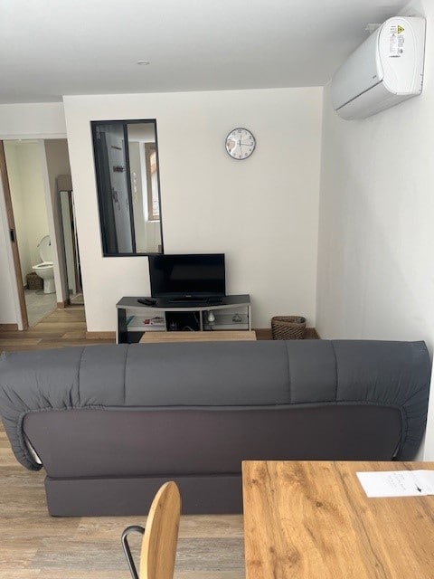 A cozy living area features a dark sofa facing a modest television stand. A round dining table is visible nearby, and a wall clock is mounted above the TV. Natural light is encouraged by nearby openings, enhancing the space's inviting ambiance.