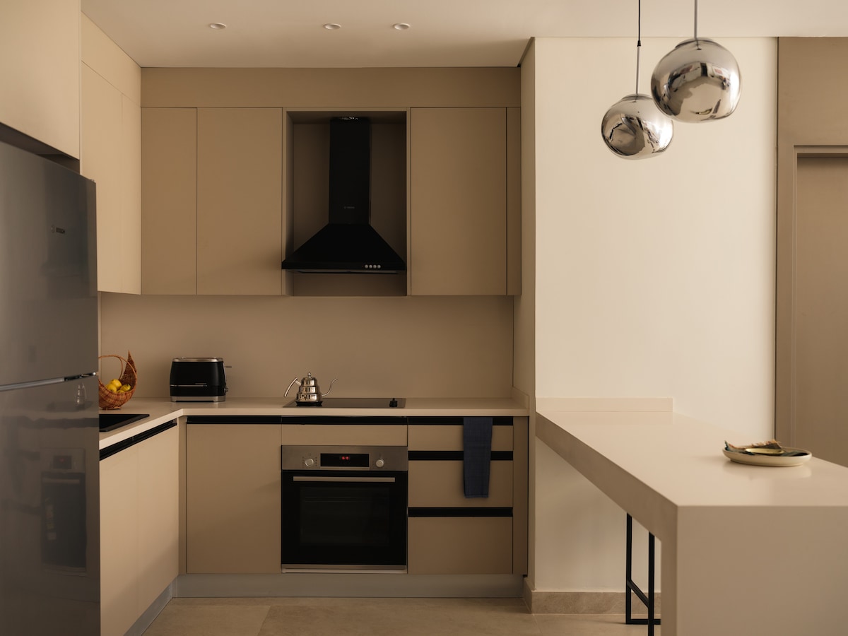 A modern kitchen is presented, featuring sleek cabinetry in neutral tones. An oven and stovetop are integrated into the minimalist design, accompanied by a stylish exhaust hood. A kitchen island offers additional counter space, and pendant lights provide functional illumination above.