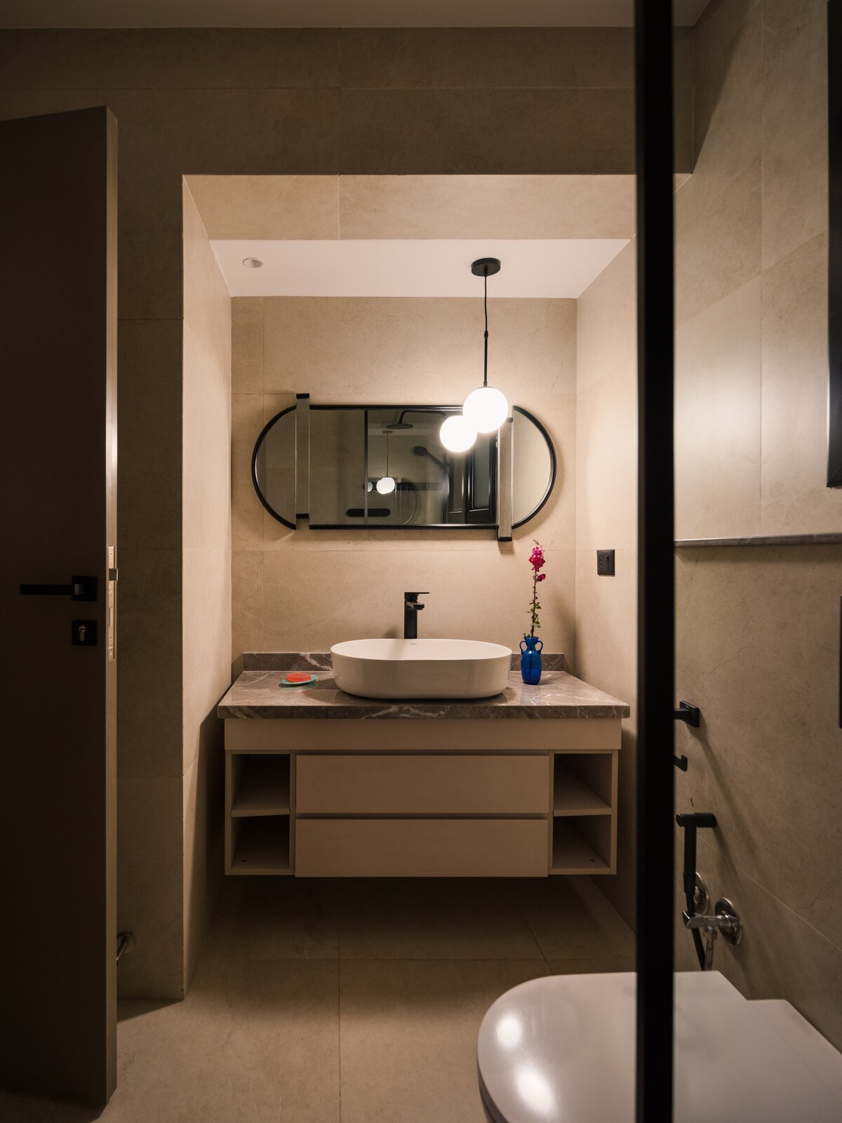 A modern bathroom features a minimalist design with neutral tones. A large mirror reflects soft lighting from a circular pendant. A white sink is positioned atop a sleek vanity with open shelving, while a small blue vase with a flower adds a subtle decorative touch.