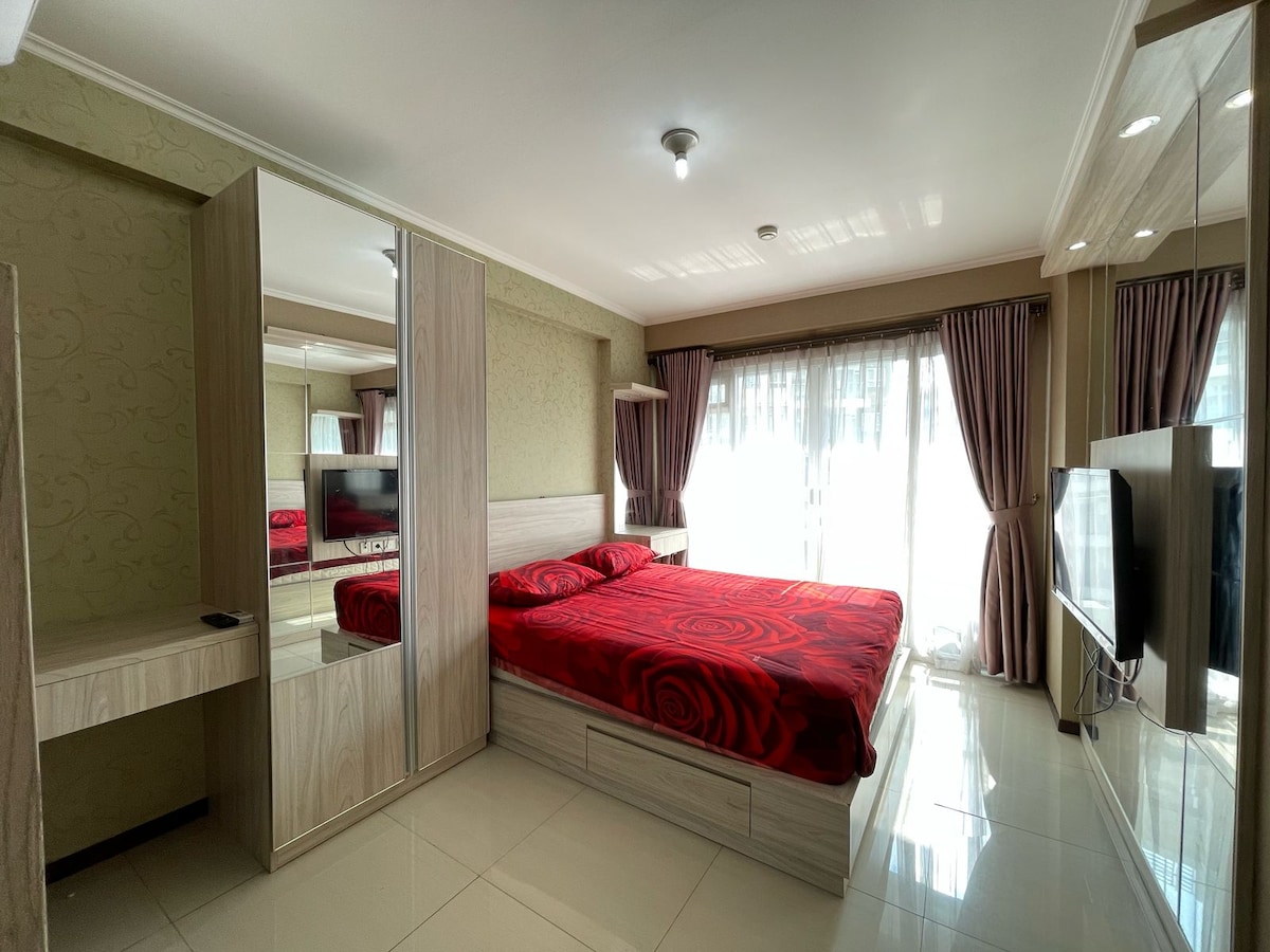 A studio apartment features a spacious bed adorned with a red rose-patterned blanket. Large mirrors reflect the room, while natural light brightens the space through glass doors leading to a balcony. Curtains frame two windows, adding to the modern design.