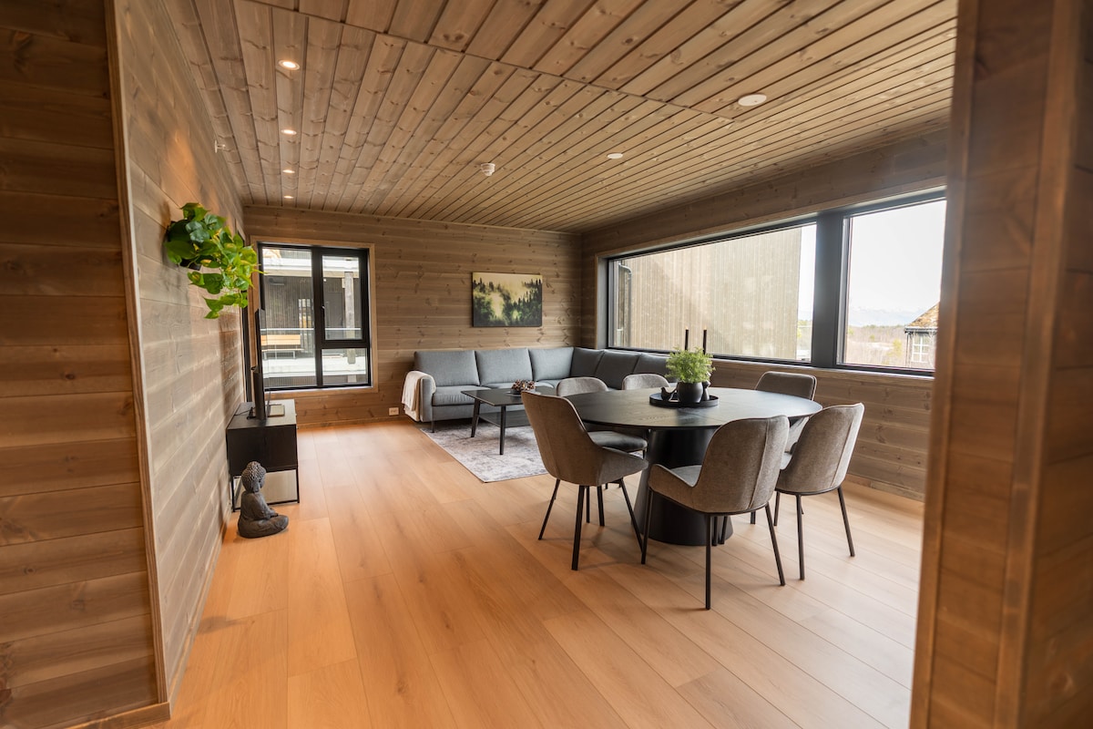 A spacious living area with wooden walls features large windows providing views of the outdoors. A round dining table is surrounded by six upholstered chairs, while a sectional sofa offers comfortable seating. Decorative plants and artwork add a touch of nature to the space.