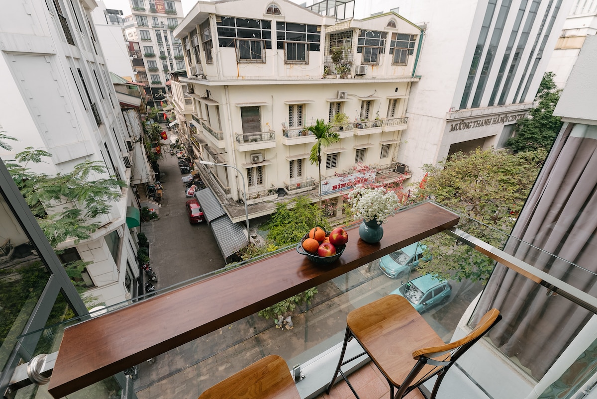 A view of the bustling Old Quarter street is captured from a glass-balustraded balcony. A wooden ledge holds a small potted plant and a bowl of fruit, enhancing the outdoor experience. Surrounding buildings reflect traditional architecture, while greenery is visible below, adding to the urban landscape.