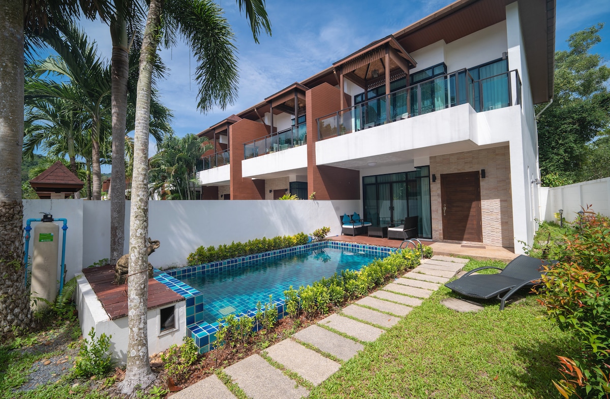 A spacious villa showcases a private swimming pool surrounded by well-manicured greenery. The property features modern architecture with large glass doors leading to a shaded patio area. Sun loungers are placed beside the pool, offering a relaxed outdoor experience.