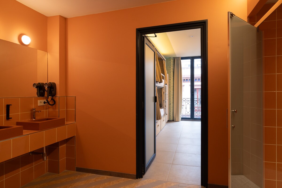 A brightly colored bathroom features orange walls and modern fixtures. Two sinks are positioned against the wall, accompanied by a wall-mounted hairdryer. A doorway leads outside, showing glimpses of a balcony accessed through sliding glass doors, revealing natural light and outdoor views.