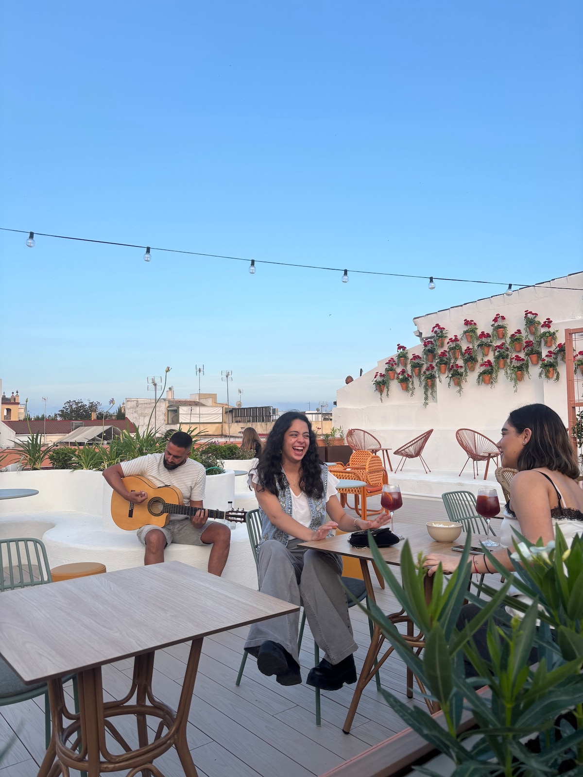 A rooftop space features several seating arrangements with stylish chairs and tables. Guests enjoy drinks while a guitarist plays nearby against a clear blue sky. Green plants and floral decorations enhance the inviting atmosphere of the terrace.
