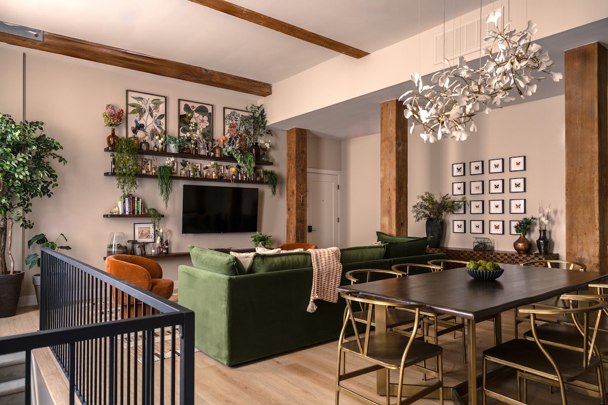 A spacious living area features a large green sectional sofa paired with a cozy orange chair. A rustic dining table sits near large windows, surrounded by stylish gold chairs. The room is adorned with decorative plants, framed artwork, and a modern chandelier above the dining table.