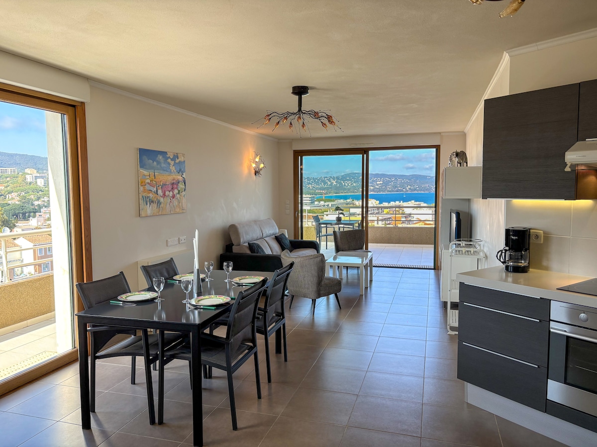 A spacious dining and living area features a glass dining table set for four, accompanied by a comfortable sofa and two armchairs. Large windows provide a panoramic view of the sea and surrounding landscape, filling the space with natural light.