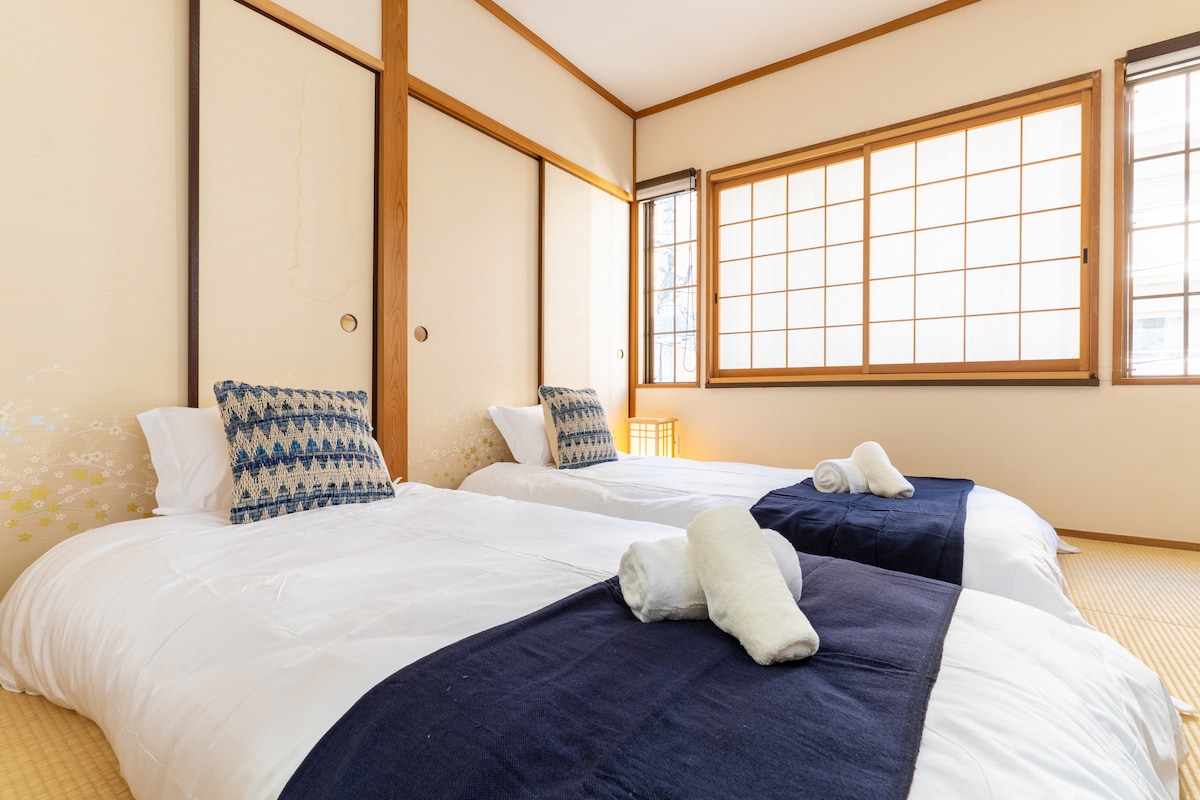 A bright bedroom is equipped with two spacious floor mattresses, each adorned with a navy blue blanket. Soft pillows are positioned atop the mattresses. Natural light filters through a large window with traditional shoji screens, illuminating the minimalist decor.