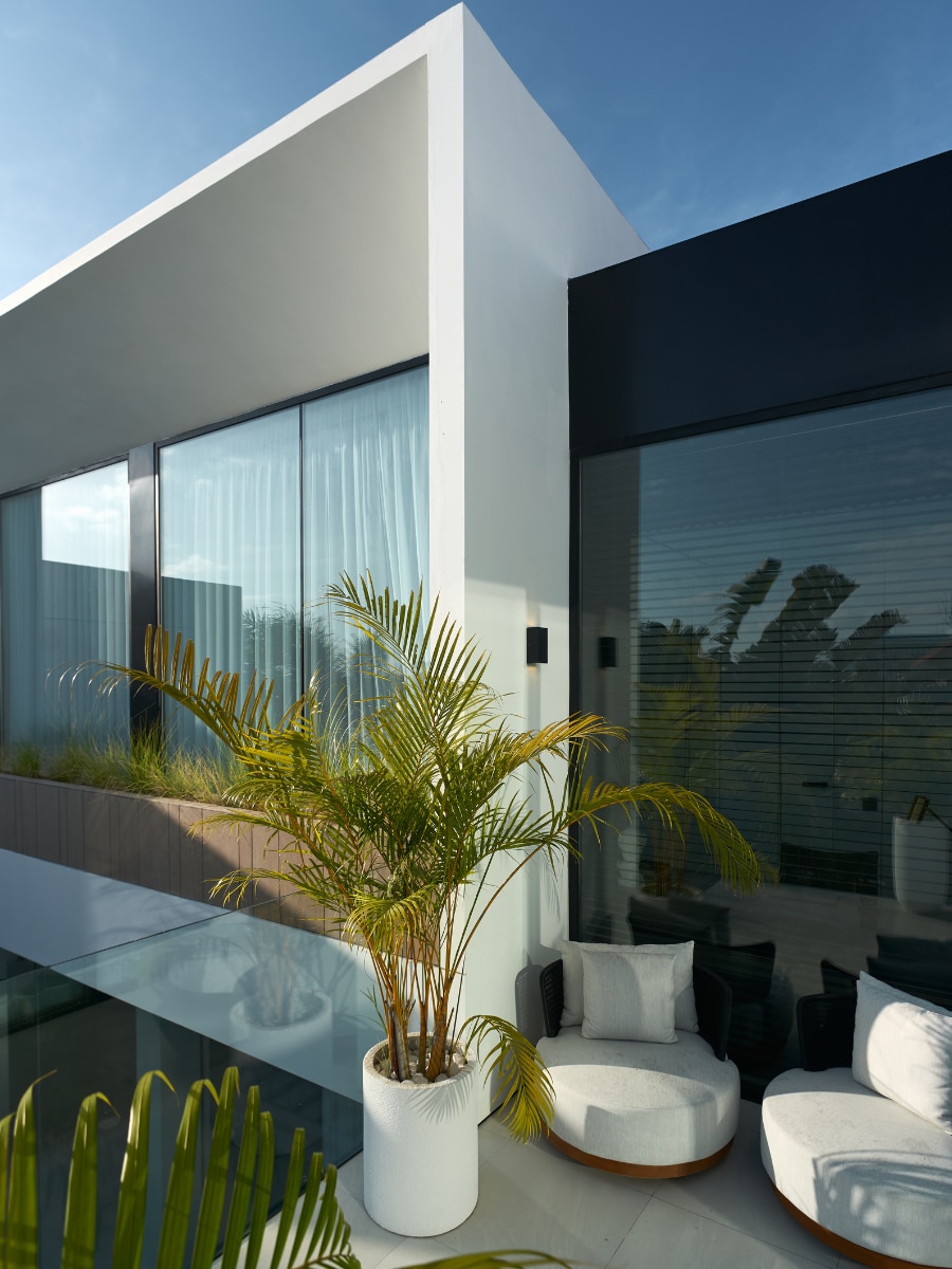 The exterior of a modern villa features sleek lines and large soundproof glass windows. A potted palm adds a touch of greenery, complementing the open seating area with comfortable cushions. The design reflects a blend of contemporary architecture and natural elements.