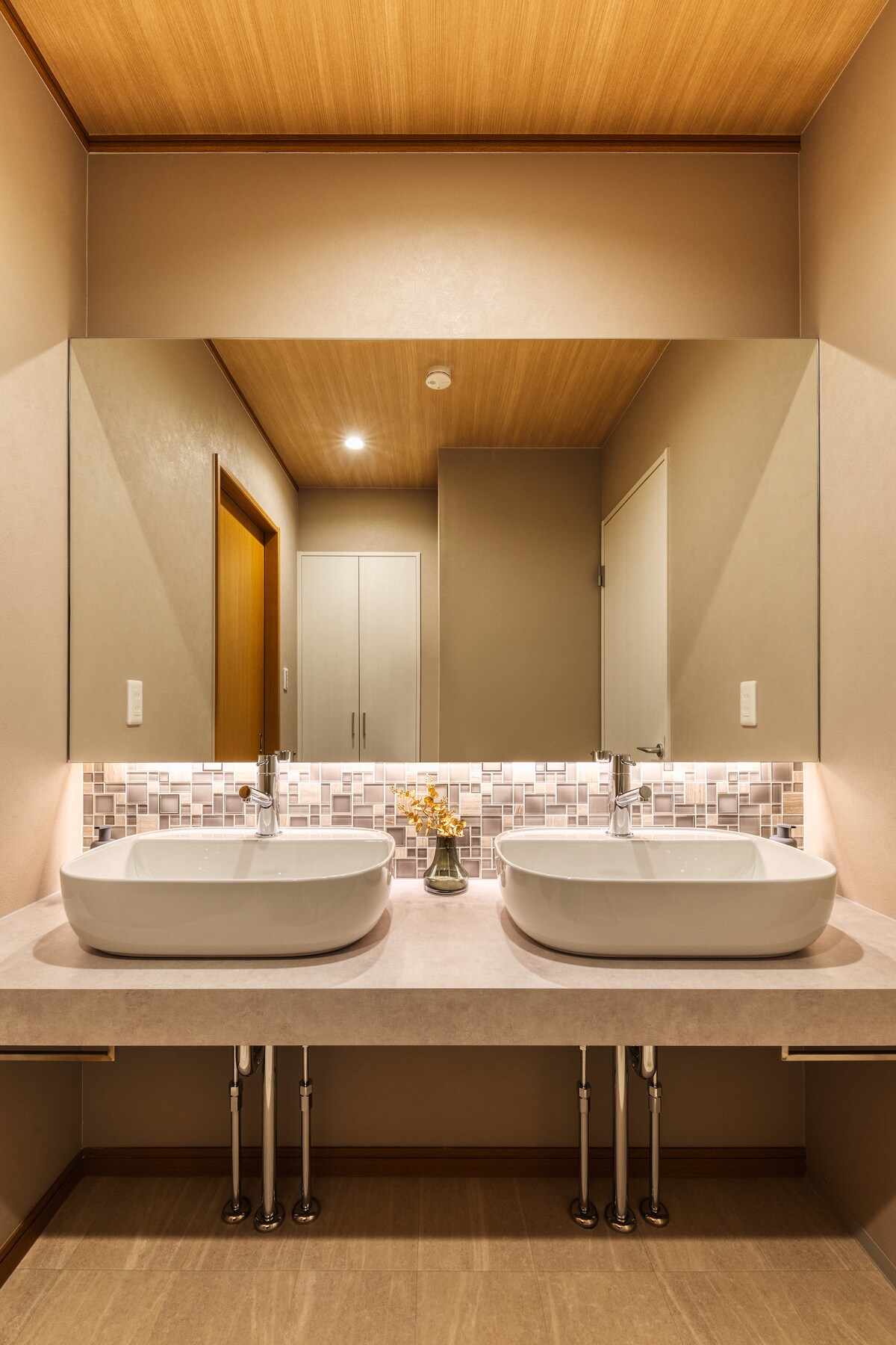 A modern bathroom is equipped with dual vessel sinks set upon a smooth countertop. A large mirror reflects the tasteful design, while soft lighting enhances the ambiance. The wall features a subtle tile backsplash, contributing to the overall clean and elegant aesthetic.