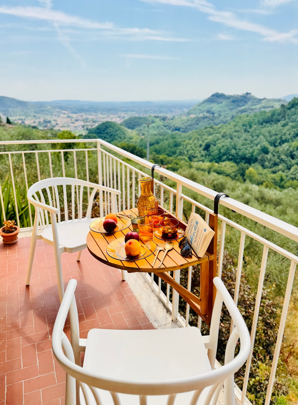 A small terrace features a wooden table adorned with a pitcher and several fruit pieces, overlooking lush green hills. Two white chairs are positioned at the table, offering a serene spot for relaxation amidst natural beauty.
