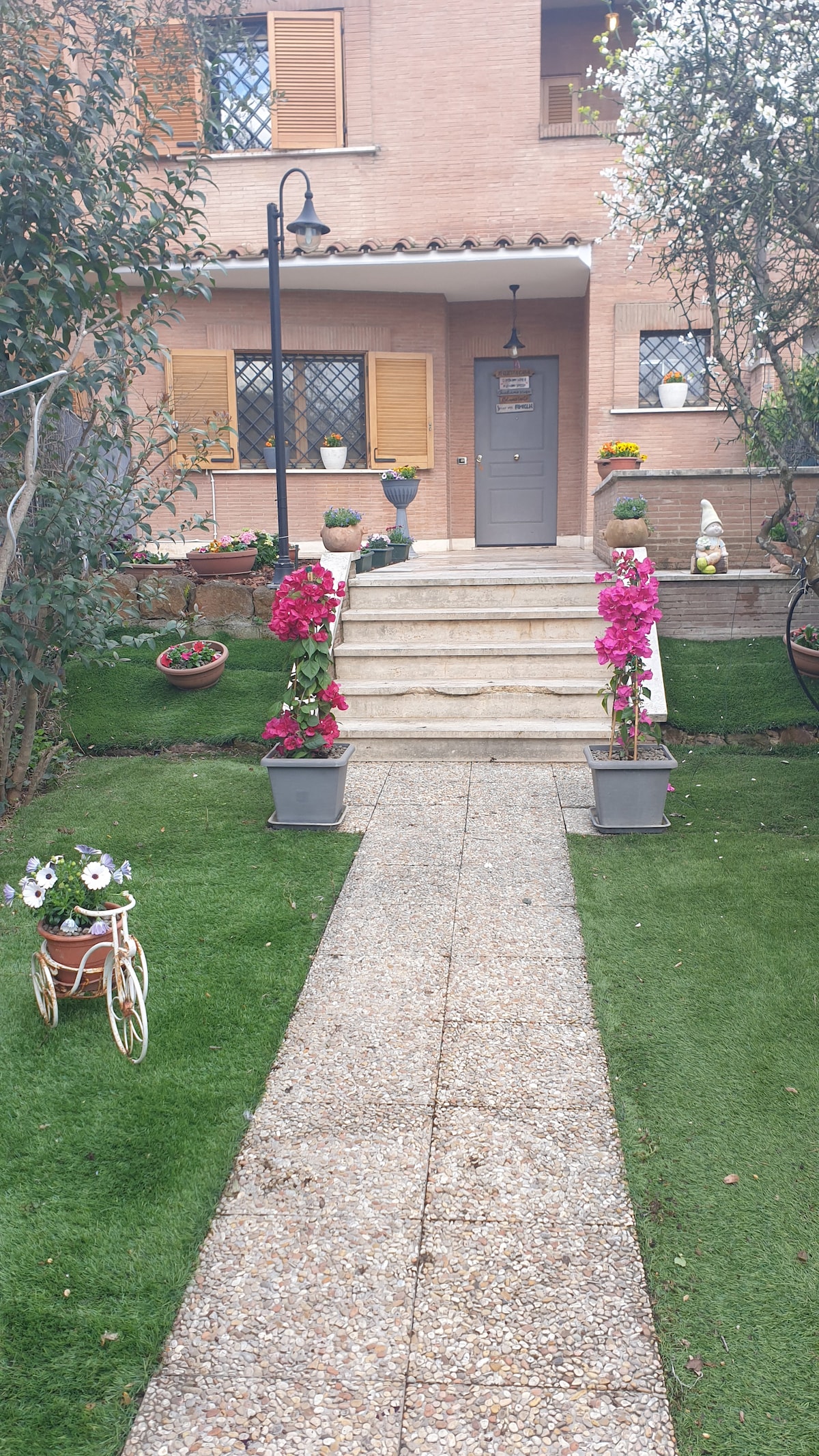 An inviting entrance is framed by blooming flower pots in vibrant pink and white. A stone pathway leads to a door with an elegant light fixture above. Lush green grass with an assortment of plants enhances the welcoming atmosphere of the property.