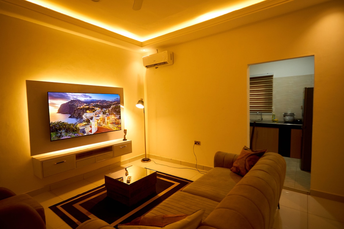 A well-lit living room showcases a large wall-mounted television displaying a scenic image. A cozy sofa is positioned around a low coffee table, with soft lighting highlighting the space. An open doorway reveals a kitchen area, adding functionality to the inviting atmosphere.