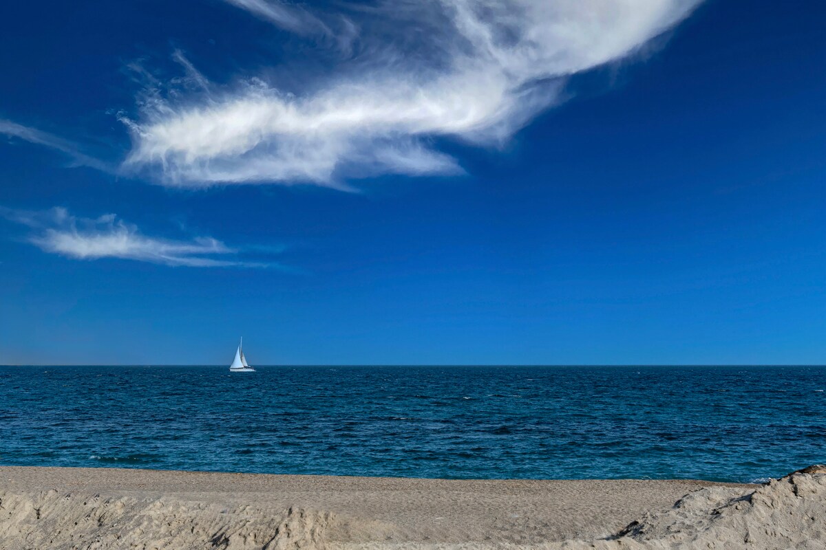 A serene coastal view features a vast expanse of the sea meeting the sky, complemented by a lone sailboat gliding calmly across the water. The sandy beach is visible in the foreground, gently curving along the shoreline under a clear blue sky.
